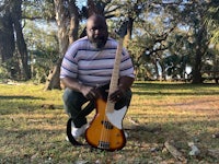 a man crouching in front of a tree with a bass guitar